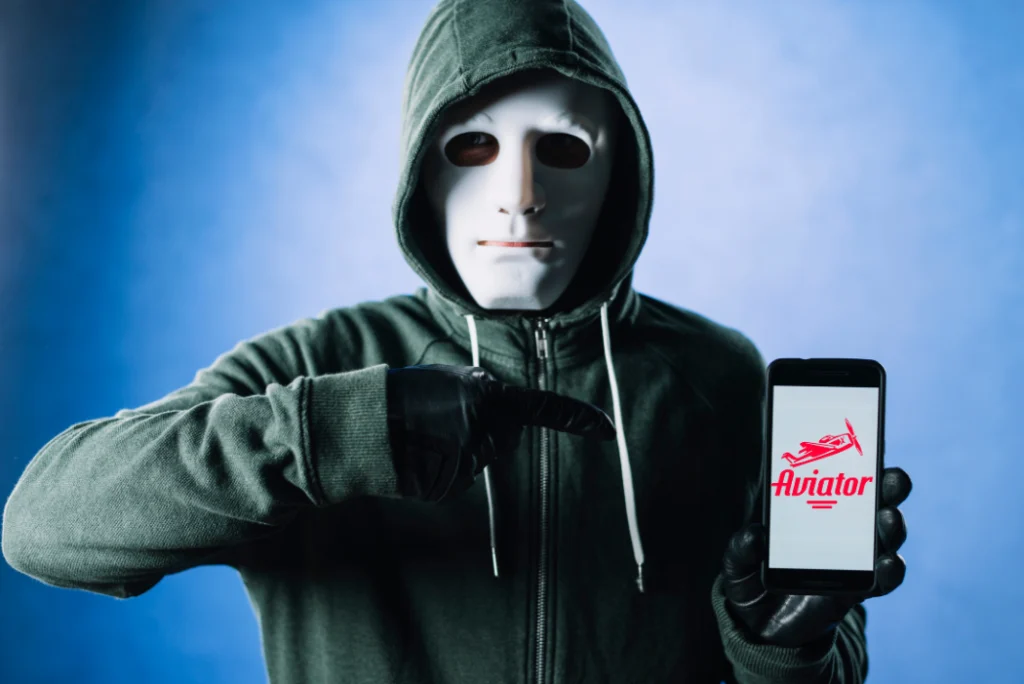 A hacker in the white mask holding smartphone which displays Aviator game logo